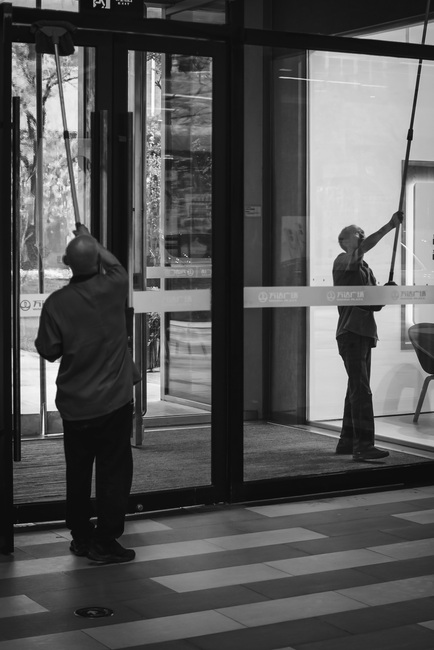 A city street scene outside a modern glass building with the logo of a retail store, featuring pedestrians and a street cleaner in reflective orange hi-vis workwear. The worker, holding a long street