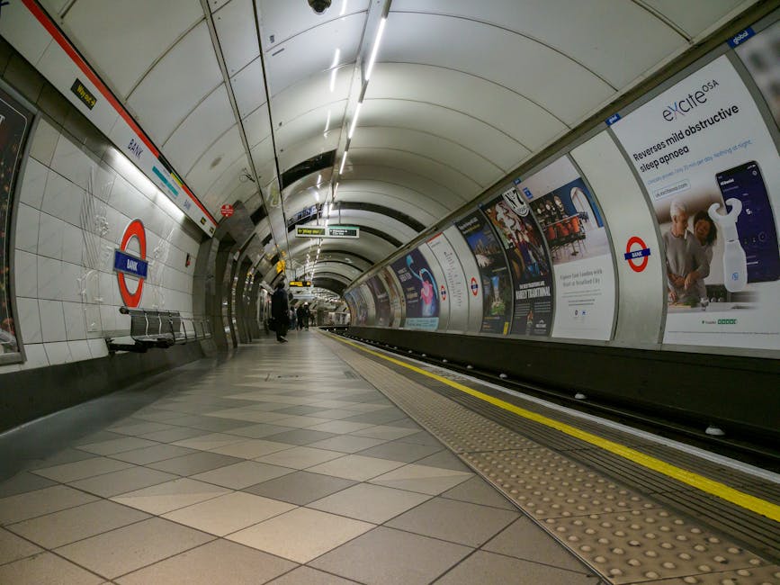Photograph of Stockwell Station platform showing painted blue metal supporting columns and beams, with signs hanging from the ceiling indicating train schedules. The platform surface is made of smooth